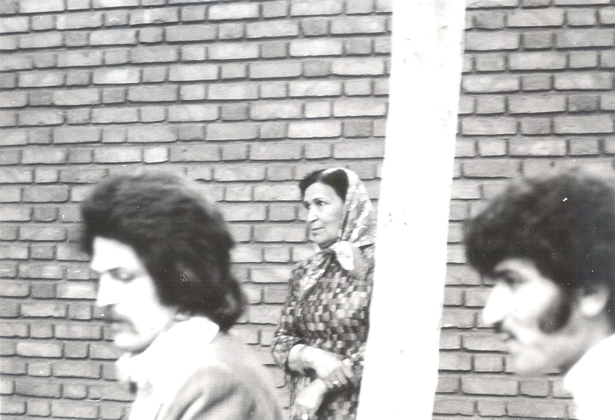 Candid street photo from 1970s Iran showing an older woman in a headscarf contrasted with modern hairstyles of younger men.