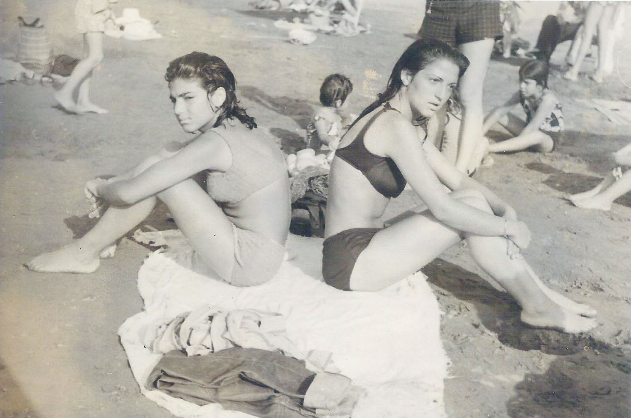 Two young women in swimsuits sitting back-to-back on a towel at a sandy beach in pre-revolutionary Iran.