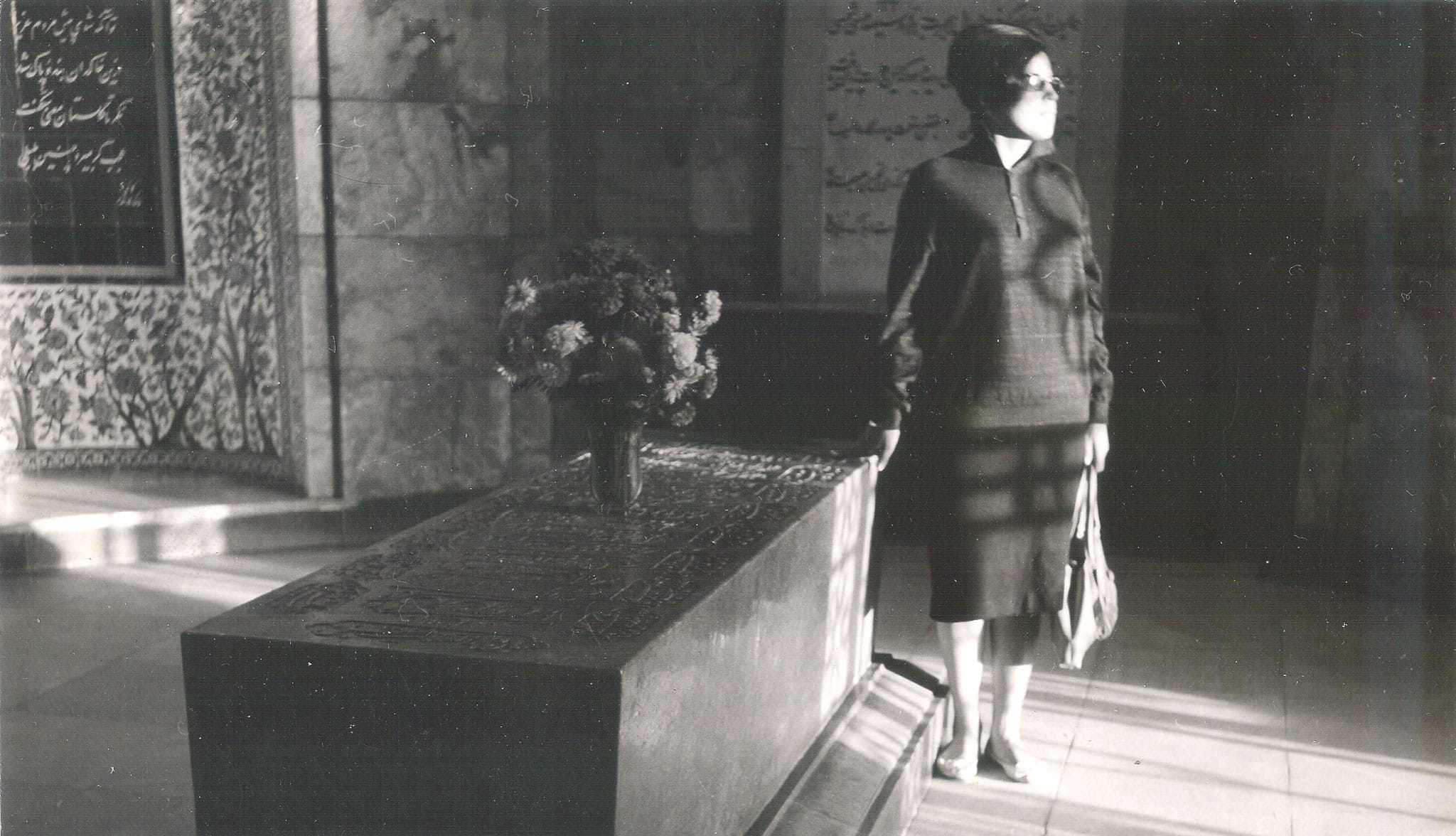 A woman in a dark dress standing solemnly beside a large engraved stone tomb inside a sunlit Iranian shrine.