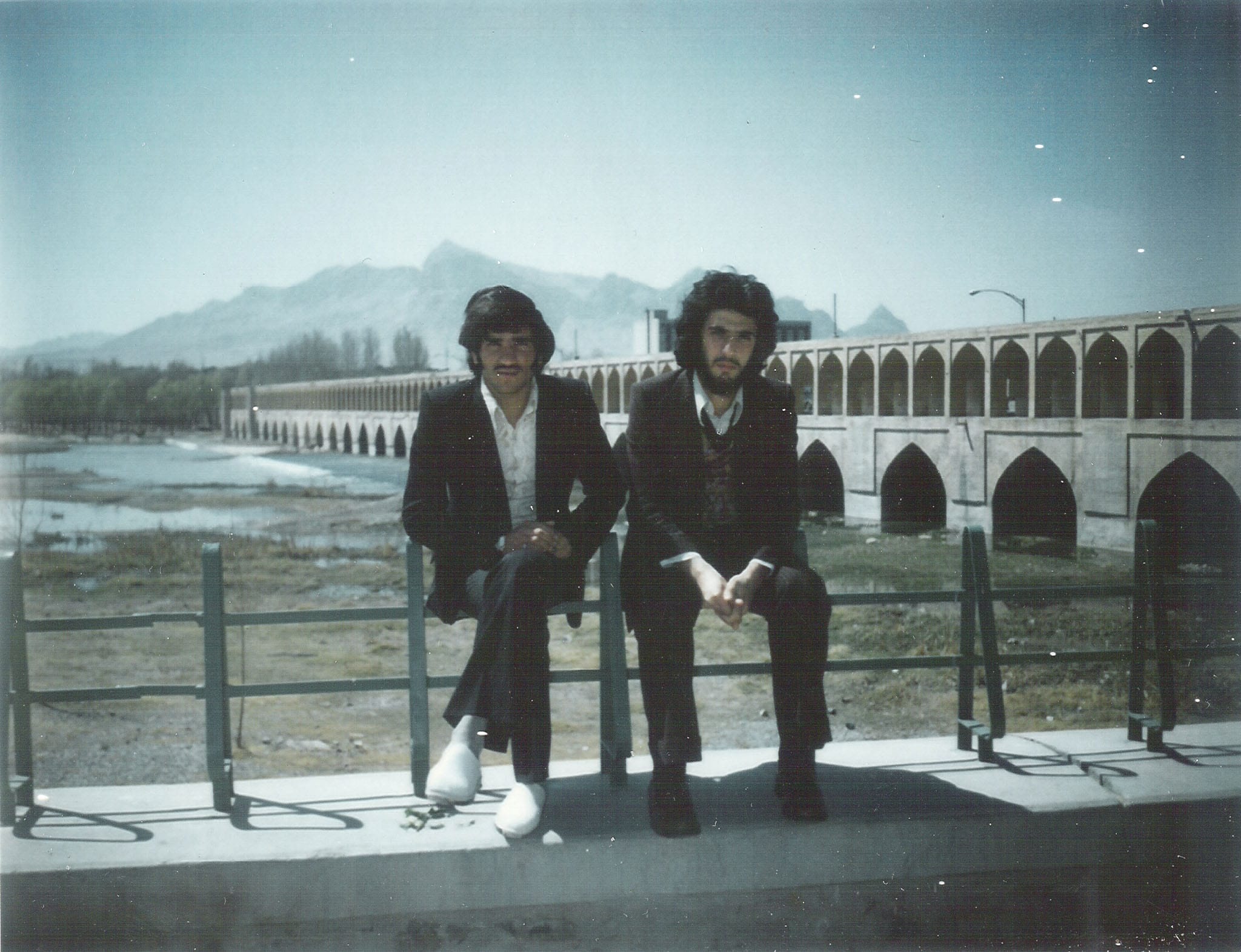 Two young men in 1970s suits and long hair sitting on a railing in front of the historic Khaju Bridge in Isfahan, Iran.