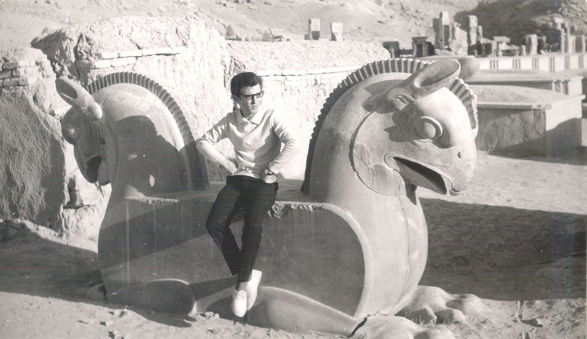 A young man wearing glasses sitting on a large ancient Homa bird stone carving at the Persepolis ruins in Iran.