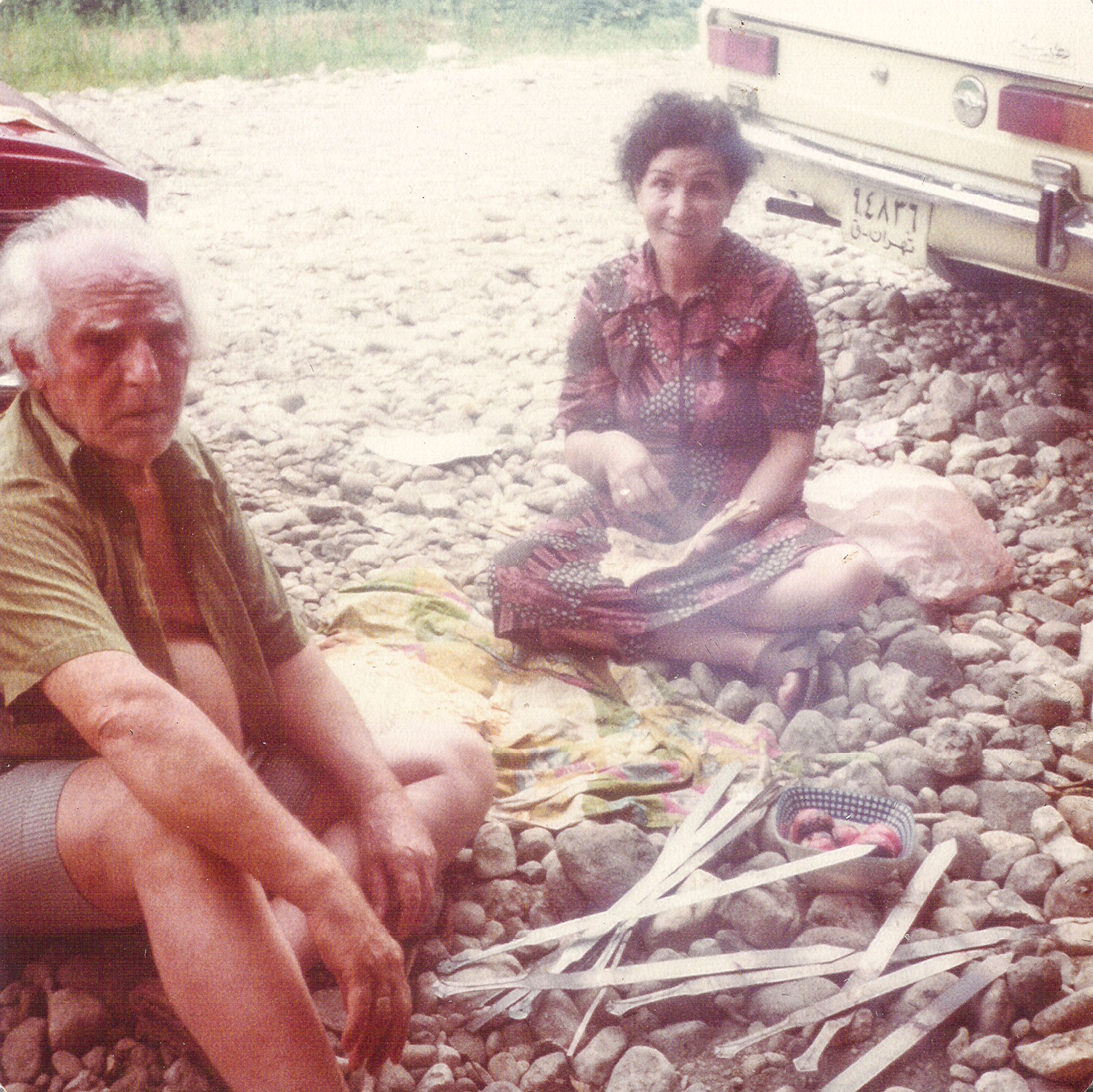 An older couple having a picnic on a rocky roadside in Iran, with metal kebab skewers and a cream-colored Paykan nearby.