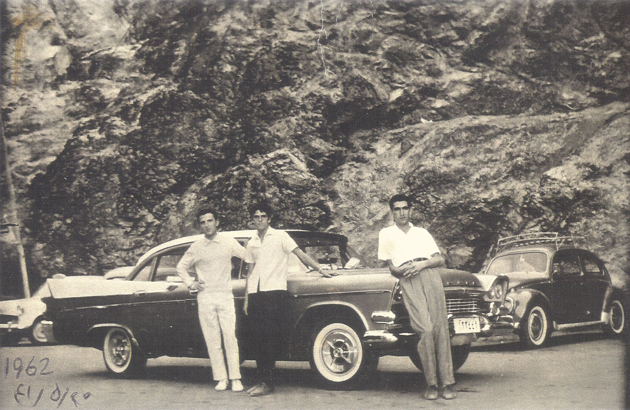 Three young men posing with a classic American car and a Volkswagen Beetle in front of a rocky mountain face.