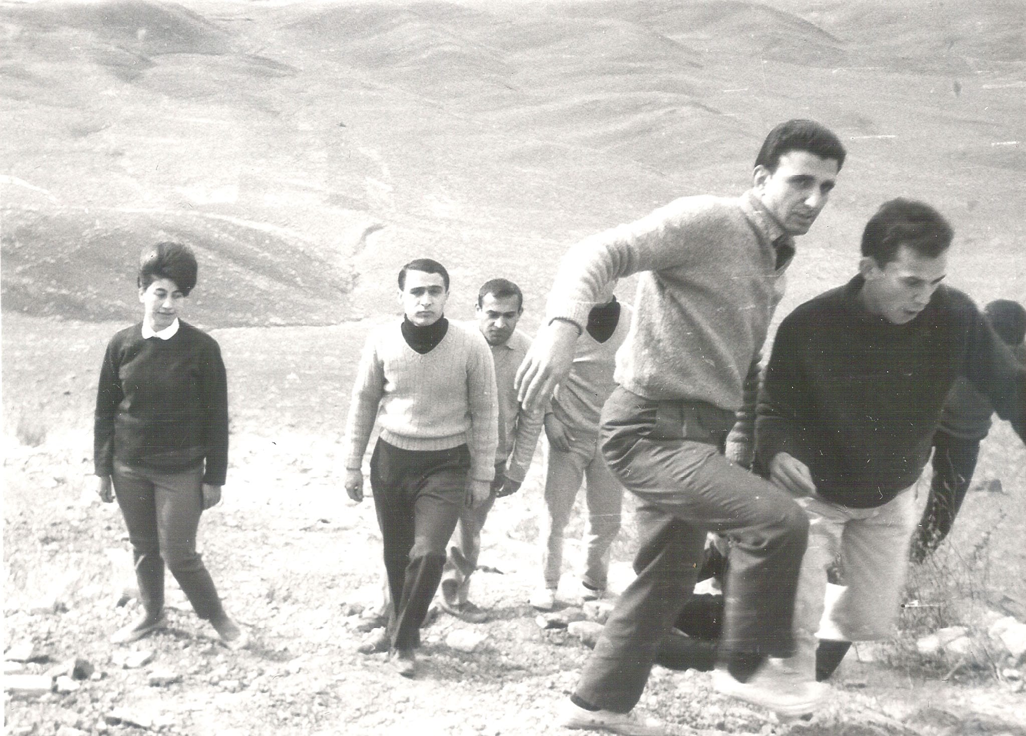 A group of male and female college students in sweaters and slacks hiking up a dry, rocky hillside.