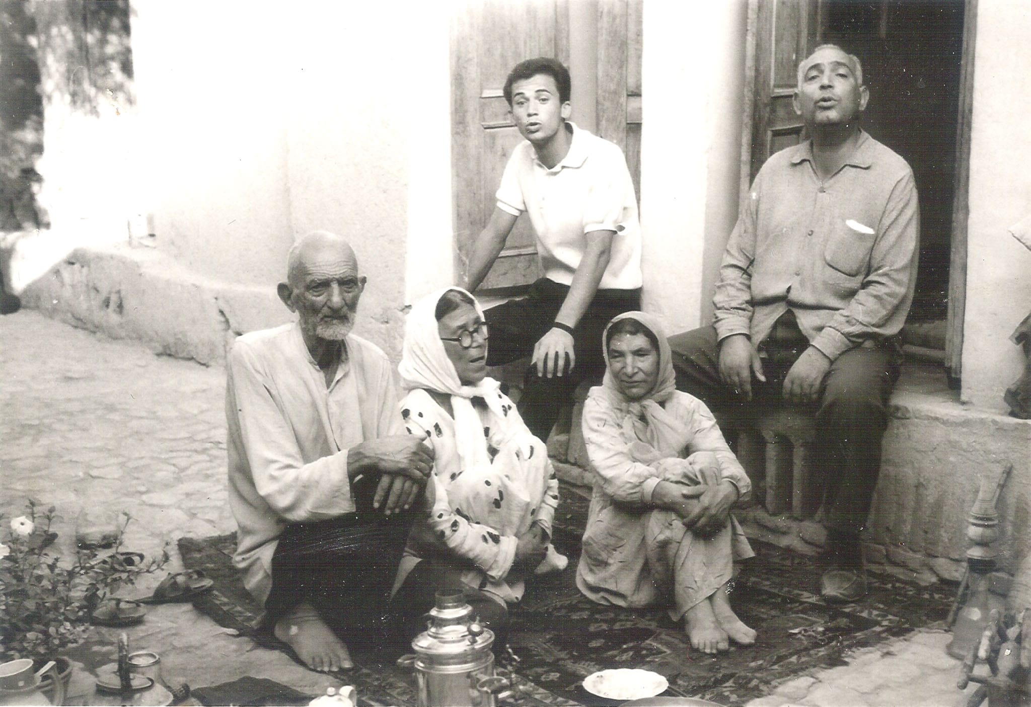 A multi-generational Iranian family sitting on a rug in a courtyard with a silver samovar in the foreground.