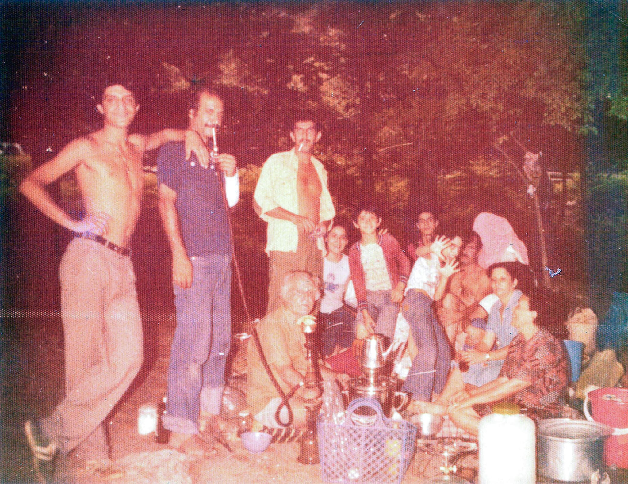 A large family gathered outdoors at night around a picnic spread, with men smoking shisha (hookah) and a traditional samovar in the center.