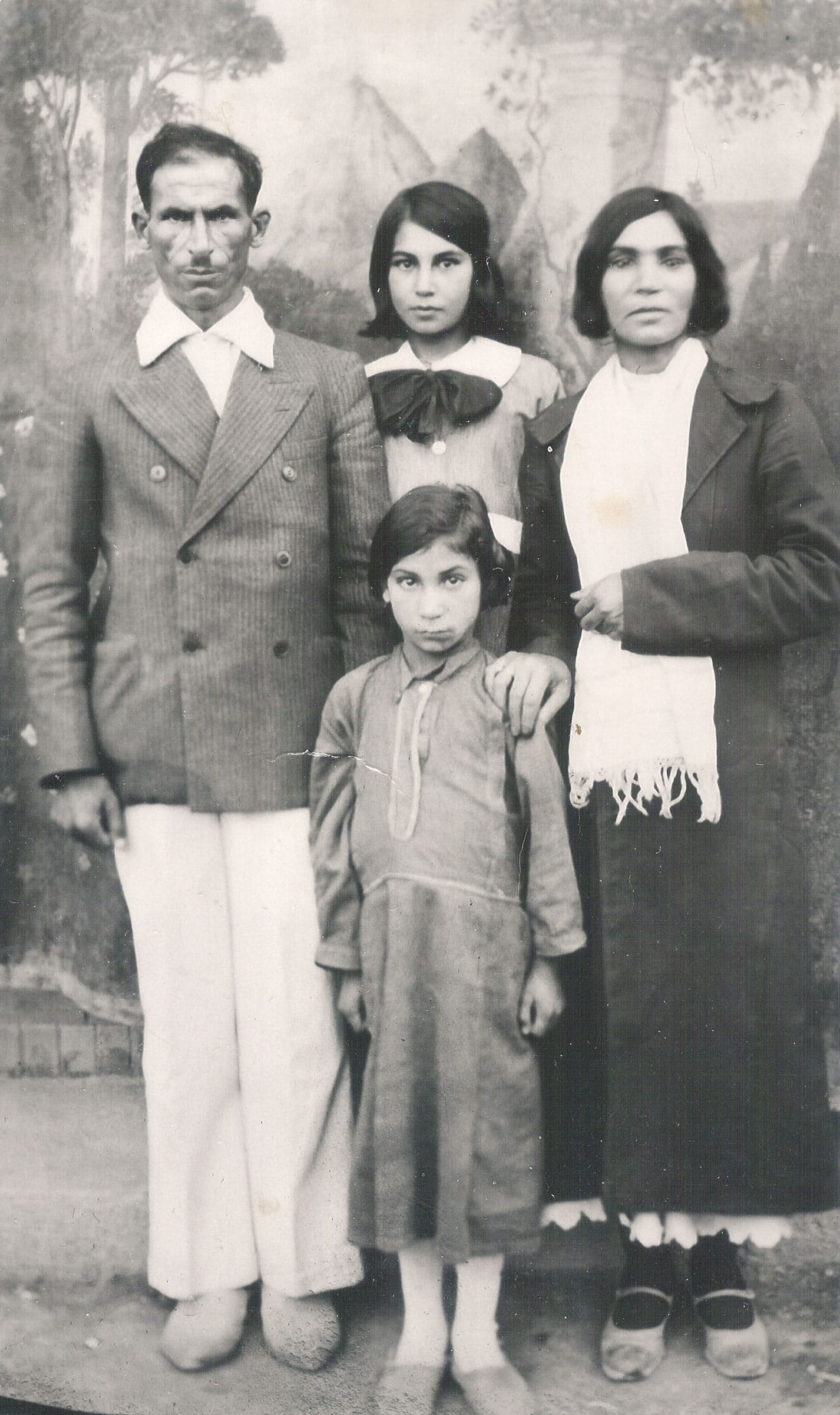 A formal black and white studio portrait of a father in a pinstripe blazer, a mother in a dark coat and white scarf, and two young daughters in dresses.