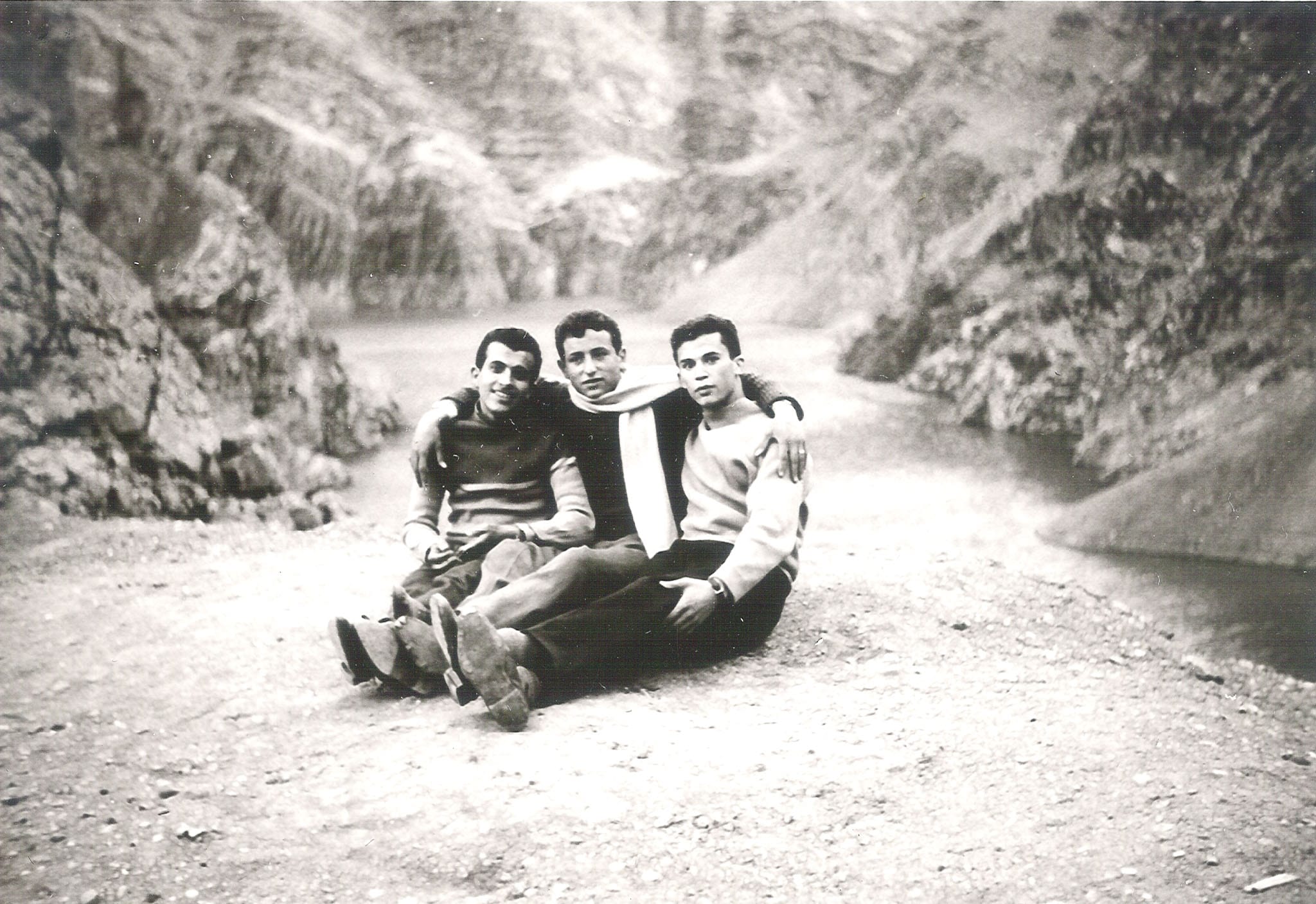 Three young men in sweaters and a scarf sitting with their arms around each other on a high mountain ridge overlooking a river or lake.