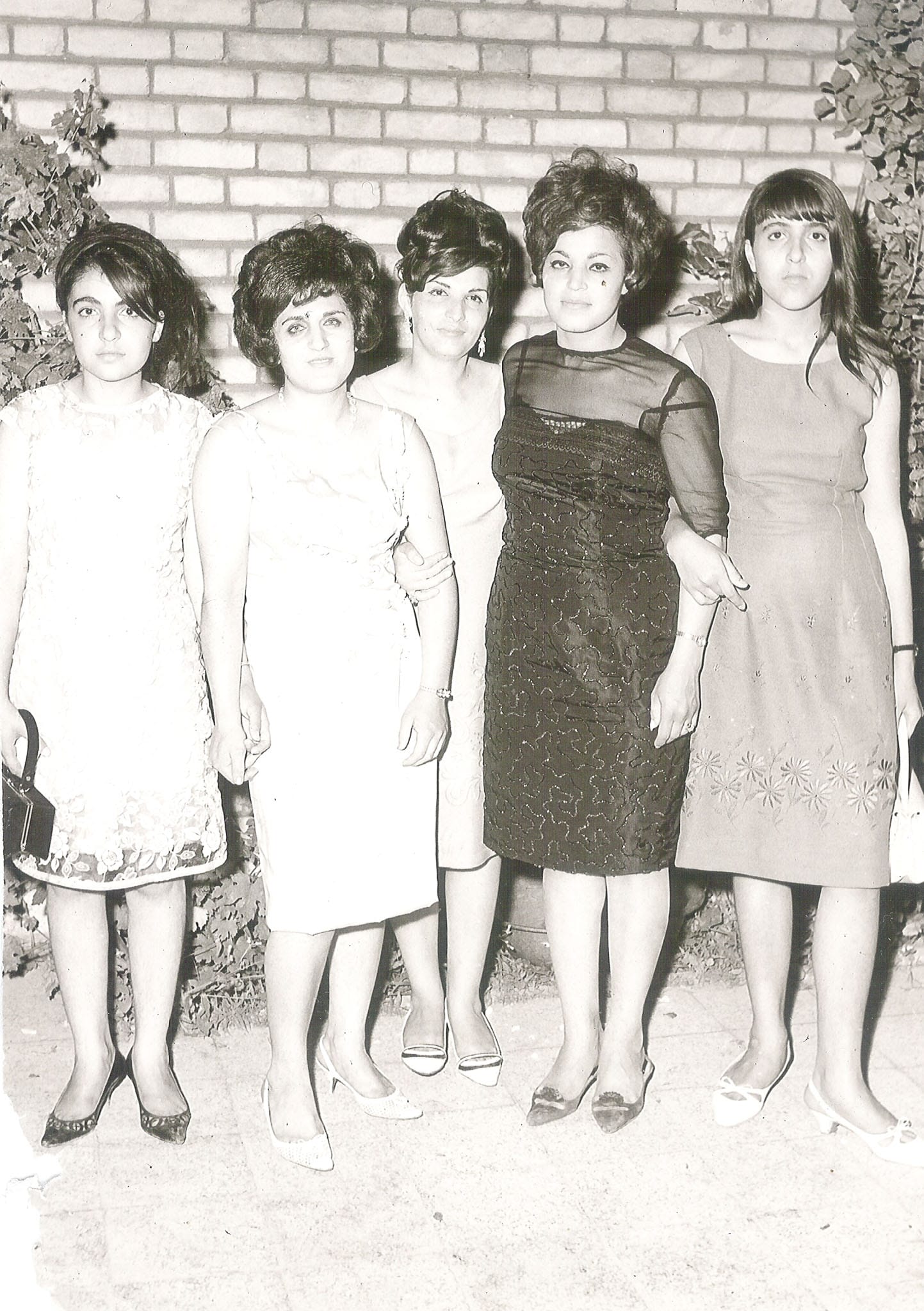 Five young women standing in a row in front of a brick wall, wearing elegant cocktail dresses and signature 1960s beehive and bouffant hairstyles.