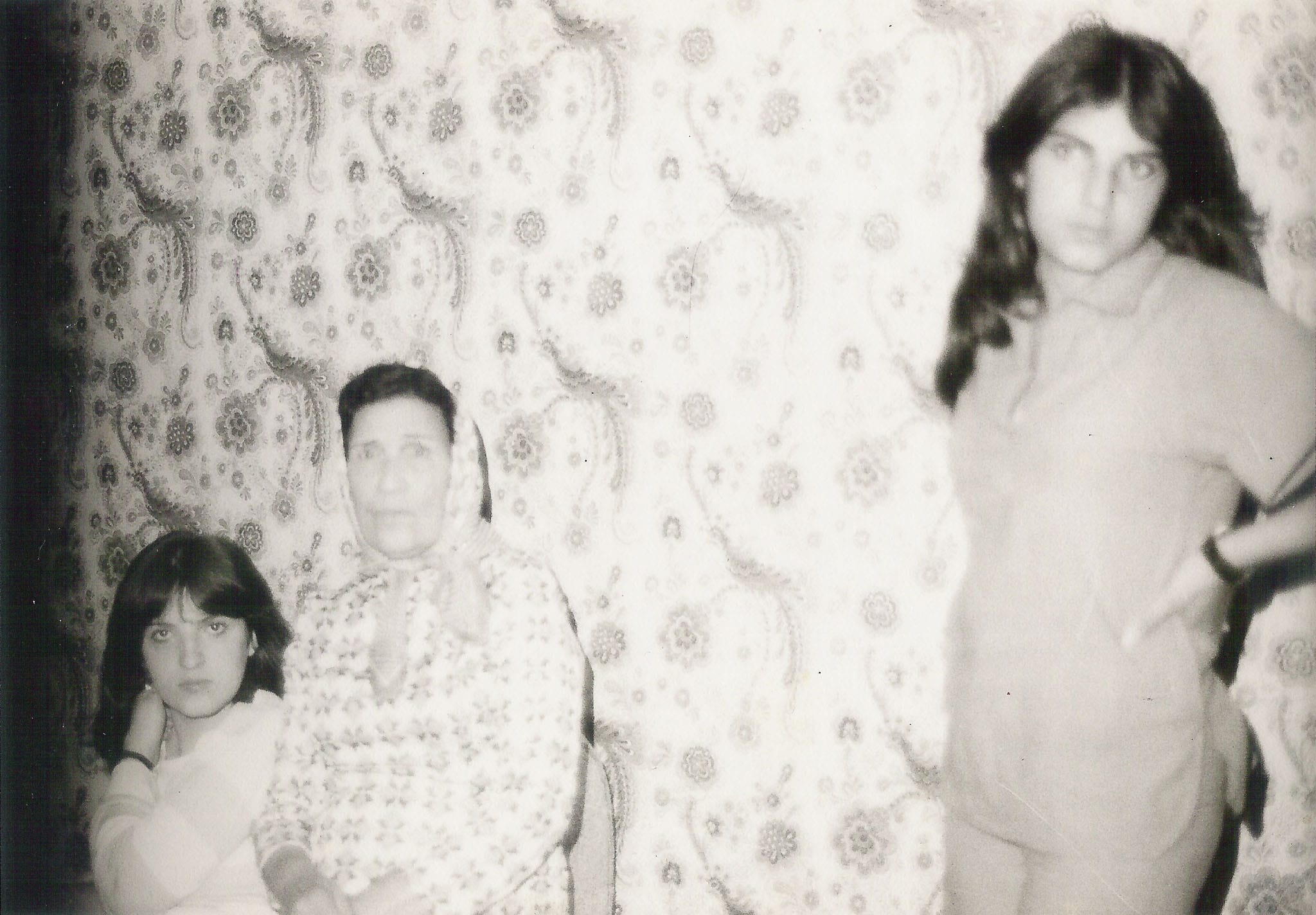 Three women, including an elder in a headscarf and a young woman in a polo shirt, posing against floral wallpaper.