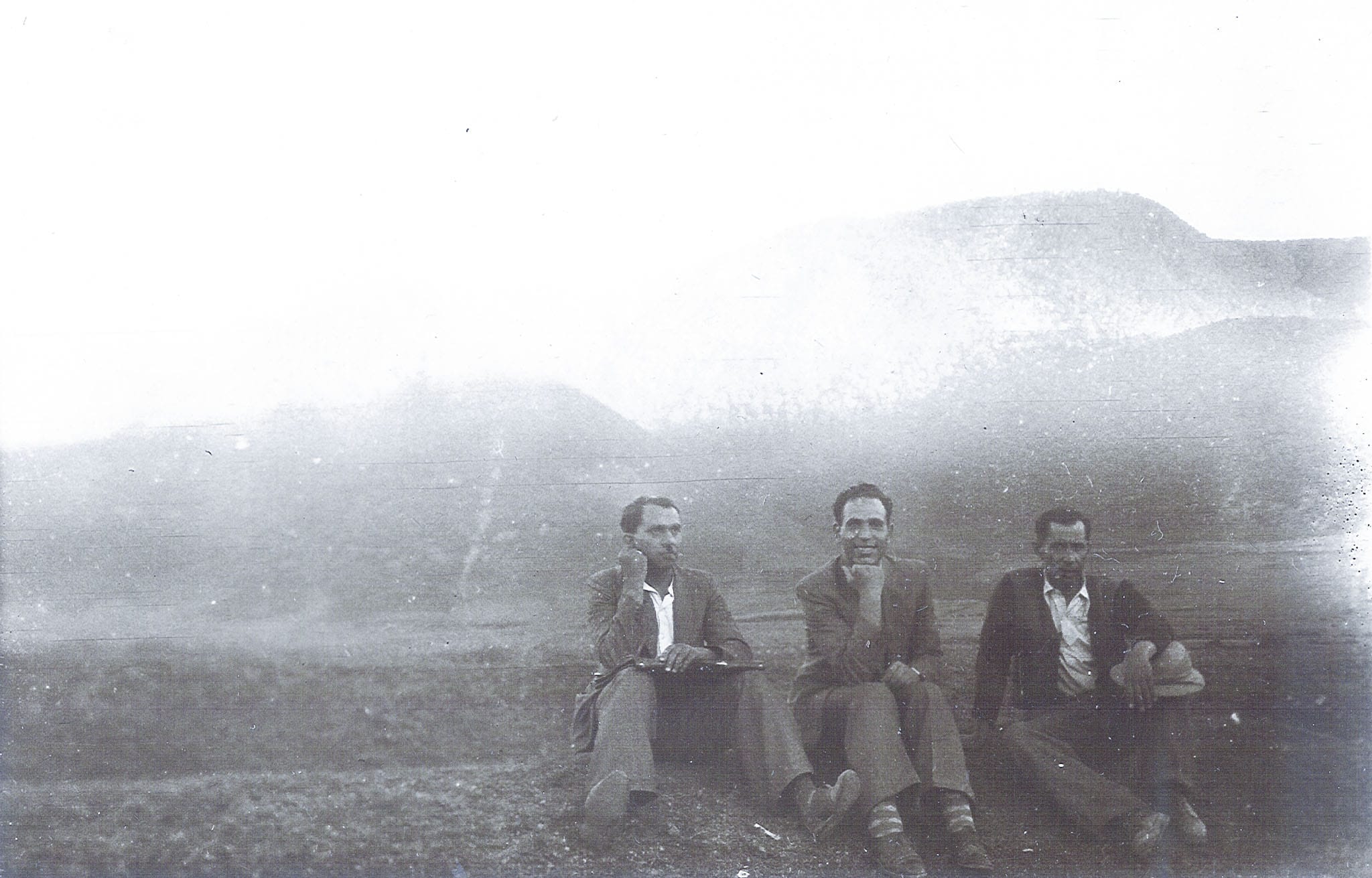 Three men in suits sitting on a grassy hilltop against a backdrop of misty, hazy mountains.