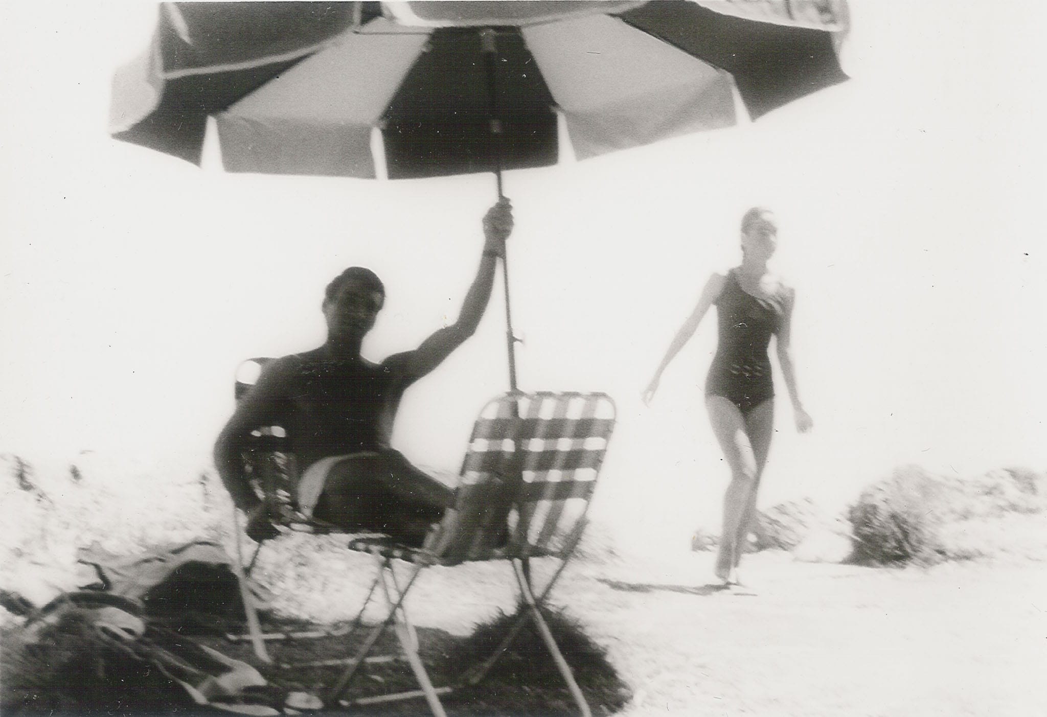 A man relaxing under a beach umbrella in a lawn chair while a woman in a swimsuit walks along the sandy shore.
